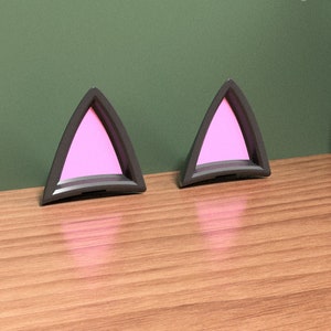 Puede incluir: Un par de orejas de gato rosas con contornos negros. Las orejas están hechas de un material suave y afelpado y son perfectas para agregar un toque de fantasía a cualquier atuendo.