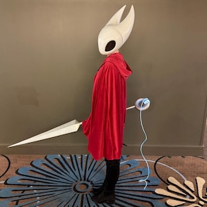 May include: A cosplay costume featuring a white helmet with large horns, a red hooded cloak, and a large white sword. The costume includes black boots and a glowing blue accessory.
