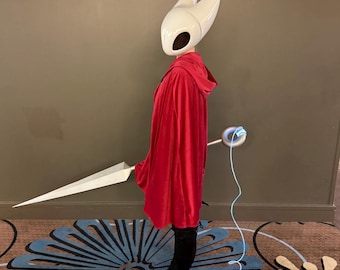 May include: A cosplay costume featuring a white helmet with large horns, a red hooded cloak, and a large white sword. The costume includes black boots and a glowing blue accessory.