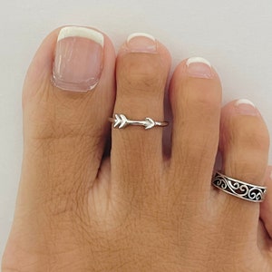 May include: Two silver toe rings. One ring features a simple arrow design. The other ring has a more intricate, swirling design.