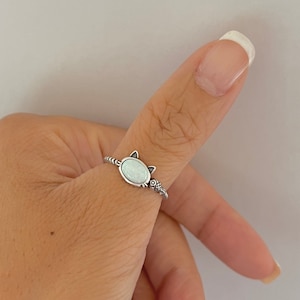 May include: A silver ring featuring a white opal stone set in the shape of a cat's head. The ring band is twisted and has a small detail on the side.