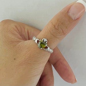 May include: A silver Claddagh ring with an olive-green heart-shaped gemstone, a small diamond, and hands clasping the heart. The ring is displayed on a finger against a white backdrop.