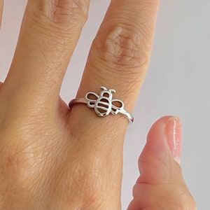 May include: A silver ring featuring a bee design. The bee has a detailed body and wings, with a simple band. The ring is worn on a finger, showcasing the delicate craftsmanship and the ring's overall design.