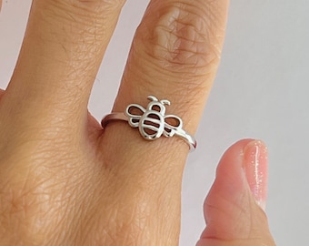 May include: A silver ring featuring a bee design. The bee has a detailed body and wings, with a simple band. The ring is worn on a finger, showcasing the delicate craftsmanship and the ring's overall design.