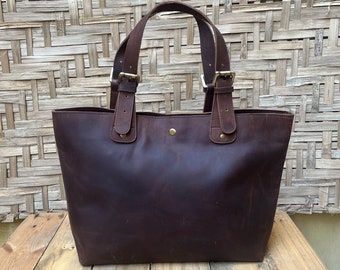 May include: A brown leather tote bag with two adjustable straps and brass buckles. The bag has a simple design and is made of high-quality leather.