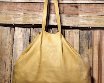 May include: A large, tan leather tote bag with long shoulder straps. The bag has a soft, slouchy design and is made of a smooth, matte leather. The bag is hanging against a rustic wooden background.