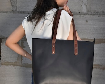May include: A black leather tote bag with brown leather straps and handles. The bag has a simple design and is large enough to carry everyday essentials.