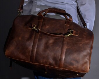 May include: A brown leather duffel bag with a long strap, worn over the shoulder of a person wearing a white shirt and blue jeans.