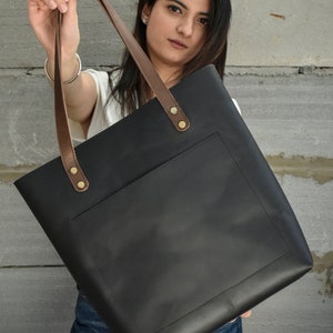May include: A black leather tote bag with brown leather straps and a front pocket. The bag has gold metal rivets on the straps.