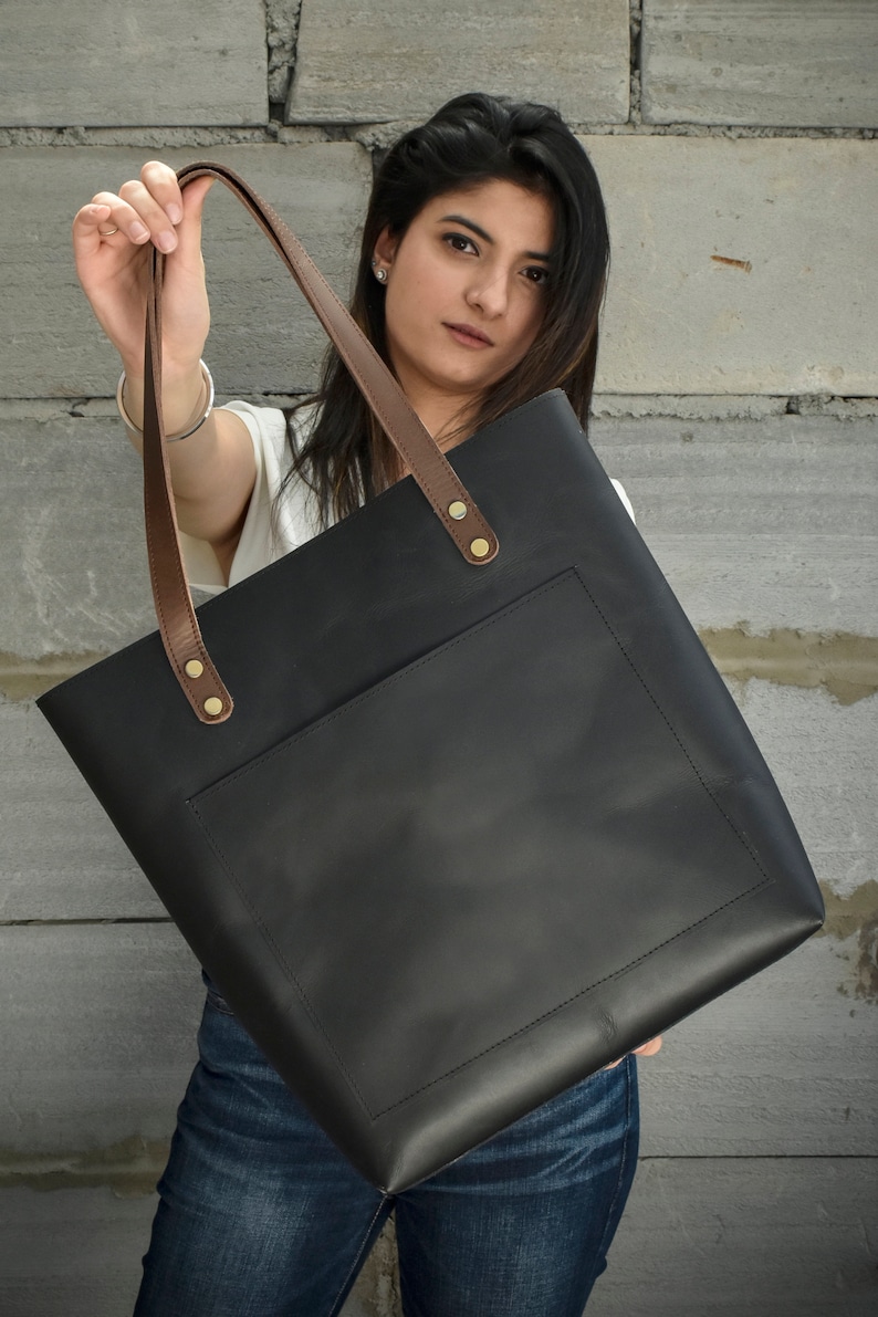 May include: A black leather tote bag with brown leather straps and a front pocket. The bag has gold metal rivets on the straps.