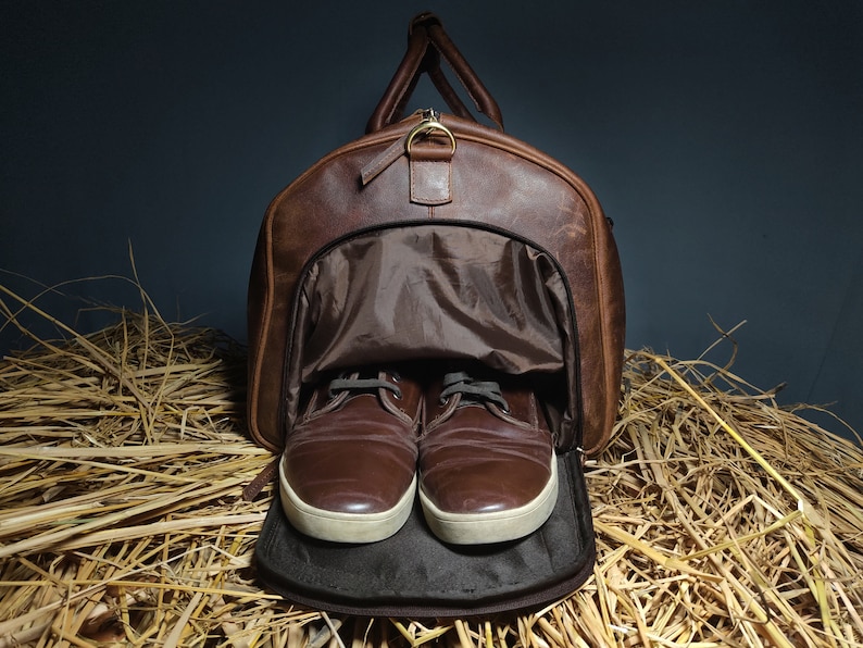 May include: A brown leather duffel bag with a zippered opening, revealing a brown fabric lining. Two brown leather shoes are inside the bag, with the laces tied. The bag is sitting on a bed of straw.