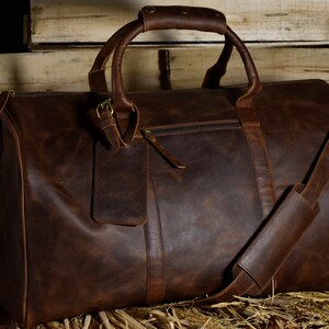 May include: A large, brown leather duffel bag with a top handle and a detachable shoulder strap. The bag has a zipper closure and a small tag attached. The leather has a distressed look.