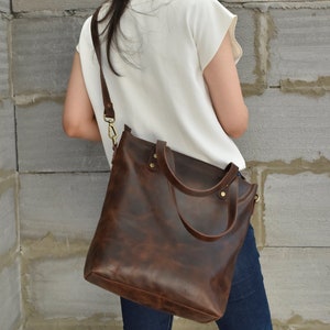 K&ouml;nnte beinhalten: Eine braune Ledertasche mit einem langen Trageriemen. Die Tasche hat einen Rei&szlig;verschluss oben und zwei obere Tragegriffe.