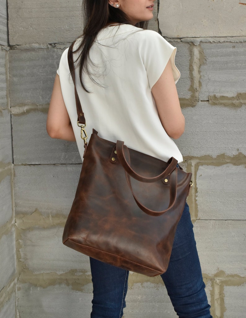 K&ouml;nnte beinhalten: Eine braune Ledertasche mit einem langen Trageriemen. Die Tasche hat einen Rei&szlig;verschluss oben und zwei obere Tragegriffe.
