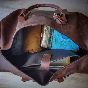 May include: Open brown leather duffel bag with a top handle and zipper closure. The bag is partially filled with items including a gold cosmetic bag, a rolled-up tan garment, and a book with the text "TOP SUMMER".