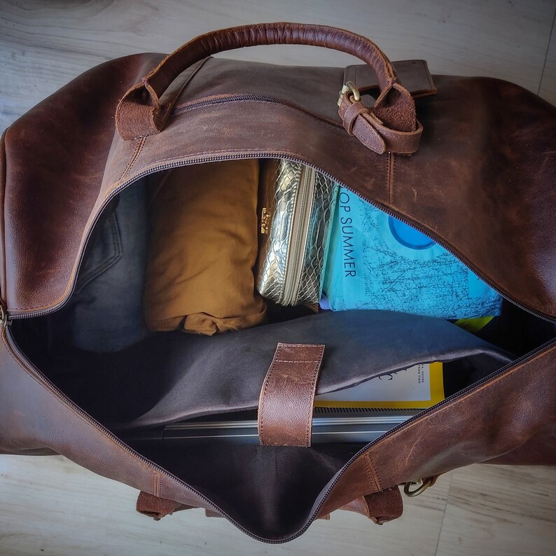 May include: Open brown leather duffel bag with a top handle and zipper closure. The bag is partially filled with items including a gold cosmetic bag, a rolled-up tan garment, and a book with the text "TOP SUMMER".