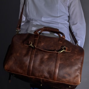 May include: A brown leather duffel bag with a shoulder strap and top handles. The bag has brass-colored hardware and is carried over the shoulder. The leather has a distressed appearance. The person is wearing a white shirt and blue jeans.