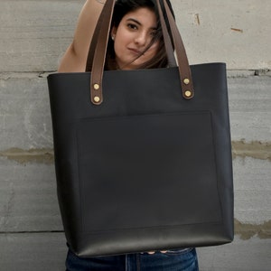 K&ouml;nnte beinhalten: Eine schwarze Ledertasche mit braunen Lederriemen und goldenen Metalldetails. Die Tasche hat eine Fronttasche und ein gro&szlig;es Hauptfach.