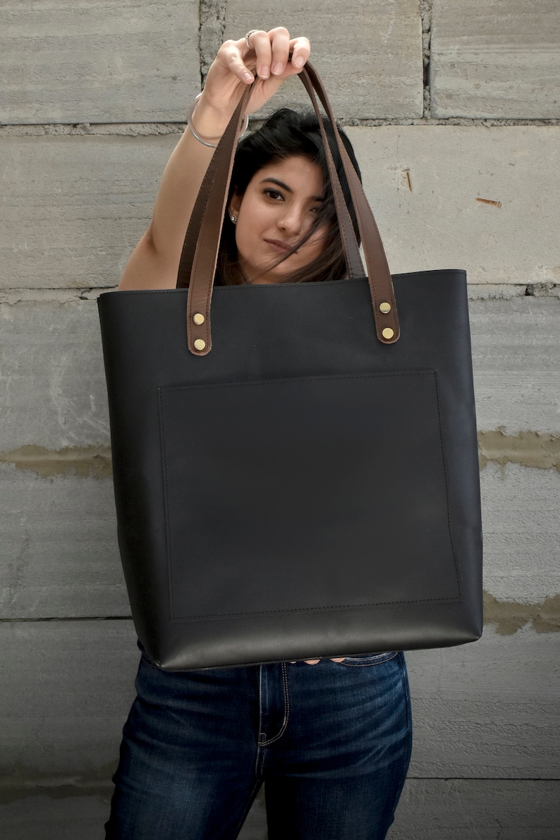 K&ouml;nnte beinhalten: Eine schwarze Ledertasche mit braunen Lederriemen und goldenen Metalldetails. Die Tasche hat eine Fronttasche und ein gro&szlig;es Hauptfach.
