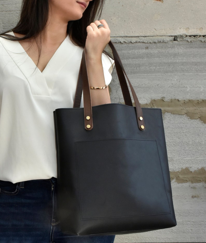 May include: A black leather tote bag with brown leather handles and a front pocket. The bag has gold-coloured metal accents.