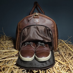 Op de afbeelding: Een bruine leren reistas met een ritsopening, die een bruine stoffen voering onthult. Twee bruine leren schoenen bevinden zich in de tas, met de veters gestrikt. De tas ligt op een bed van stro.