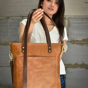 K&ouml;nnte beinhalten: Eine braune Ledertasche mit einem braunen Schultergurt und einer Fronttasche. Die Tasche hat goldfarbene Metallbeschl&auml;ge und wird von einer Person getragen, die ein wei&szlig;es Hemd und eine blaue Jeans tr&auml;gt.