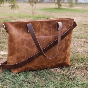 May include: A brown leather tote bag with a long strap and a front pocket. The bag has a distressed look and is made of high-quality leather.