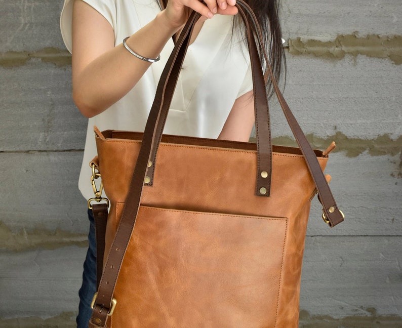 K&ouml;nnte beinhalten: Eine braune Ledertasche mit einem langen Schultergurt und zwei k&uuml;rzeren Henkeln. Die Tasche hat eine Fronttasche und einen Rei&szlig;verschluss.
