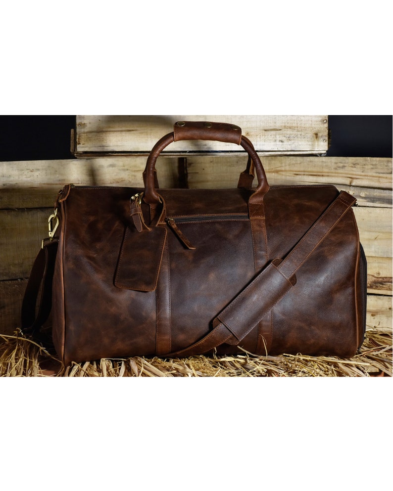 May include: A large, brown leather duffel bag with a shoulder strap and top handles. The bag has a zipper closure and a small tag attached. The leather has a distressed look, and the bag appears to be sitting on a bed of straw.