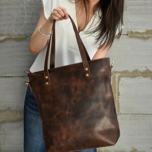 K&ouml;nnte beinhalten: Eine gro&szlig;e braune Ledertasche mit Rei&szlig;verschluss und Messingbeschl&auml;gen. Die Tasche hat einen Used-Look und wird von einer Person gehalten.