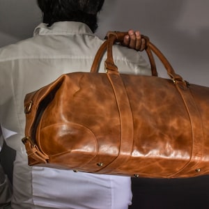 Pu&ograve; includere: Una borsa da viaggio in pelle marrone con accessori in ottone. La borsa ha una maniglia superiore e una tracolla.
