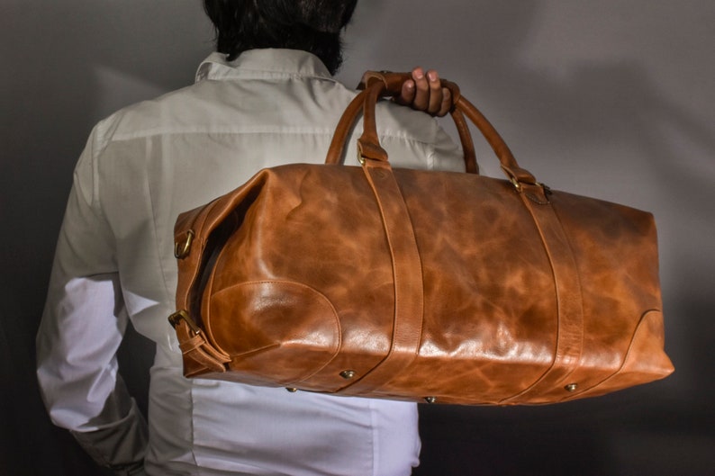 Pu&ograve; includere: Una borsa da viaggio in pelle marrone con accessori in ottone. La borsa ha una maniglia superiore e una tracolla.