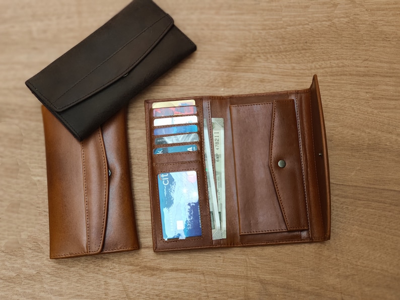 Puede incluir: Dos carteras de cuero, una marr&oacute;n y otra negra, est&aacute;n abiertas y dispuestas sobre una superficie de madera. La cartera marr&oacute;n tiene m&uacute;ltiples ranuras para tarjetas y un portafolios, mientras que la cartera negra tiene un dise&ntilde;o m&aacute;s simple.