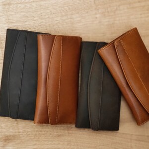 Puede incluir: Cuatro carteras de cuero en diferentes colores: negro, marr&oacute;n, marr&oacute;n oscuro y marr&oacute;n claro. Las carteras est&aacute;n cerradas y dispuestas en fila sobre una superficie de madera.