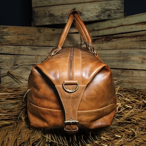 Pu&ograve; includere: Una borsa da viaggio in pelle marrone con chiusura a zip e una fibbia. La borsa ha due manici superiori e un anello a D in ottone sul davanti.