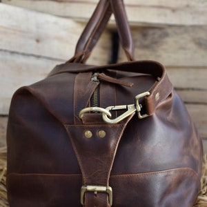 Pu&ograve; includere: Una borsa da viaggio in pelle marrone con una fibbia in ottone e una chiusura a gancio in ottone. La borsa ha un aspetto vissuto ed &egrave; realizzata in pelle di alta qualit&agrave;.