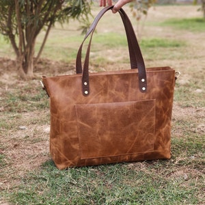 May include: A brown leather tote bag with a front pocket and brown leather straps. The bag is sitting on a bed of green grass.