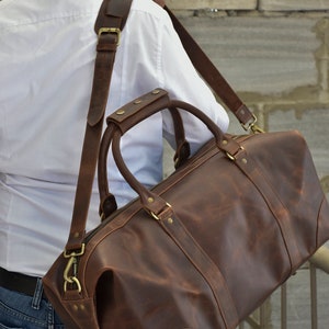 Pu&ograve; includere: Una borsa da viaggio in pelle marrone con una lunga tracolla e accessori in ottone. La borsa ha una chiusura a zip e due manici superiori.