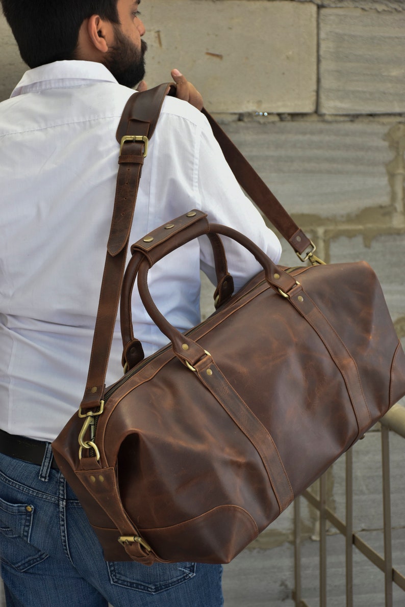 Pu&ograve; includere: Una borsa da viaggio in pelle marrone con una lunga tracolla e accessori in ottone. La borsa ha una chiusura a zip e due manici superiori.