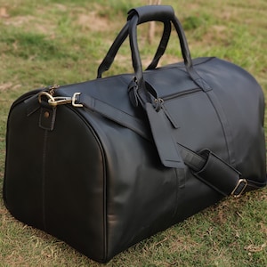 May include: A black leather duffel bag with two top handles and a shoulder strap. The bag has a zipper closure and a brass buckle.