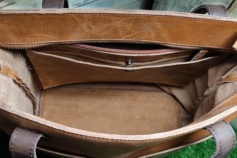K&ouml;nnte beinhalten: Das Innere einer braunen Ledertasche mit Rei&szlig;verschlussfach und braunem Stofffutter.