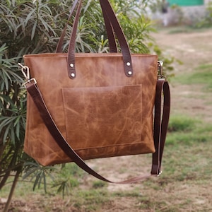May include: A brown leather tote bag with a long strap and a front pocket. The bag has a distressed look and is made of high-quality leather.