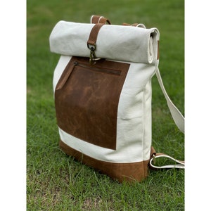 Może przedstawiać: Biały plecak z płótna z brązową klapą skórzaną i brązową skórzaną kieszenią. Plecak ma zamykanie na rolowanie i regulowane paski.