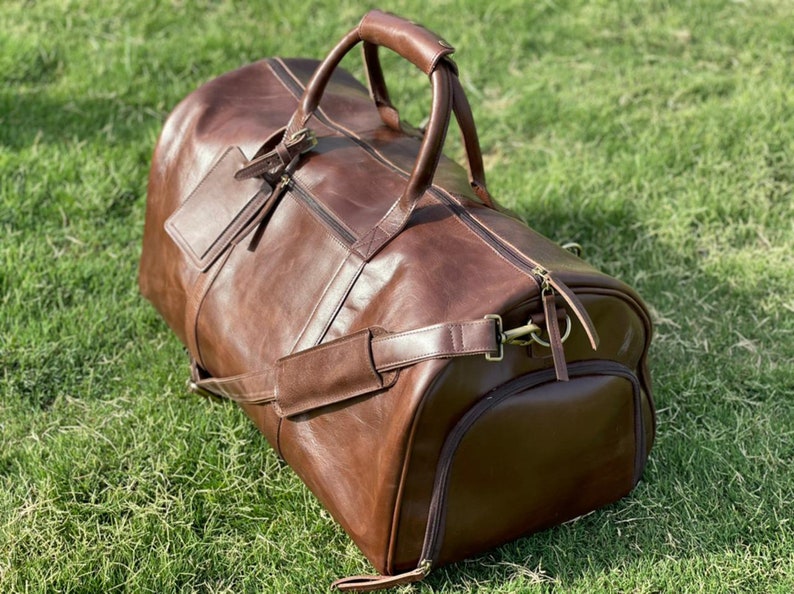 May include: A brown leather duffel bag with a top handle and a shoulder strap. The bag has a zipper closure and a small pocket on the front.