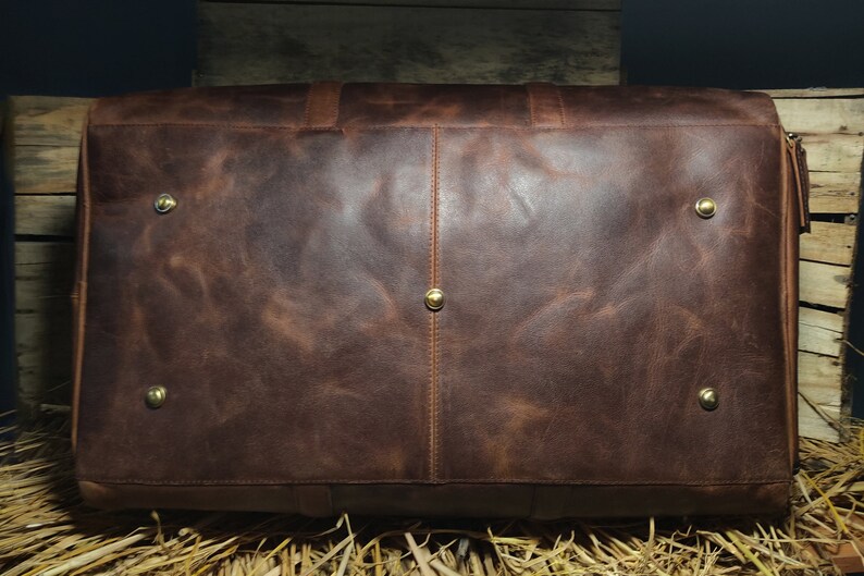 May include: A brown leather travel bag with brass-colored studs on the bottom. The bag has a handle on top and a zipper closure. The leather has a distressed look with variations in color.