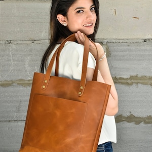 May include: A brown leather tote bag with a front pocket and gold accents. The bag is being held by a person wearing a white shirt and blue jeans.