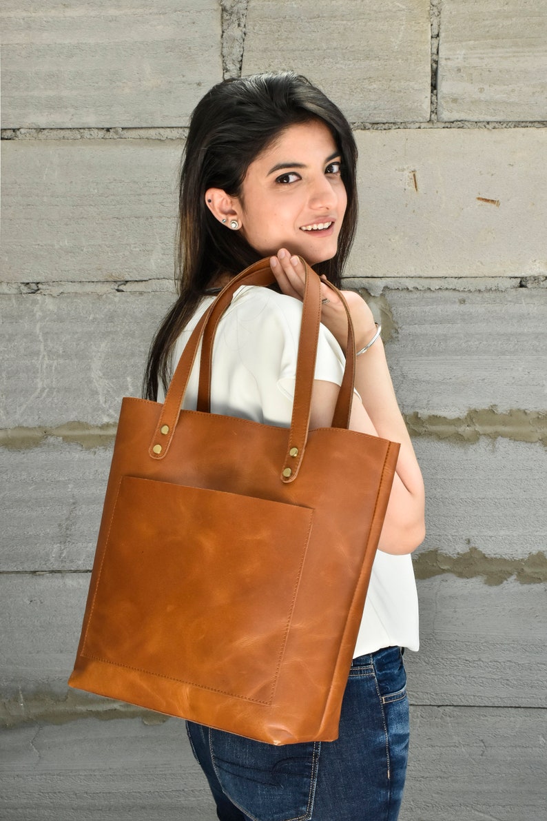 May include: A brown leather tote bag with a front pocket and gold accents. The bag is being held by a person wearing a white shirt and blue jeans.