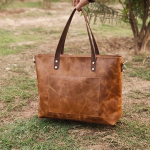 May include: A brown leather tote bag with a front pocket and brown leather handles. The bag is sitting on a bed of green grass.