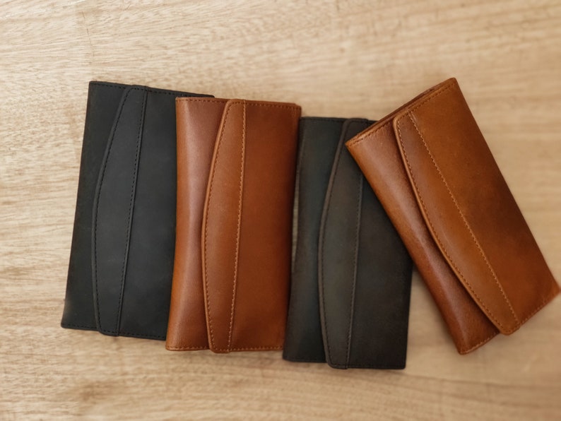 May include: Four leather wallets in different colors: black, brown, dark brown, and light brown. The wallets are closed and laid out in a row on a wooden surface.
