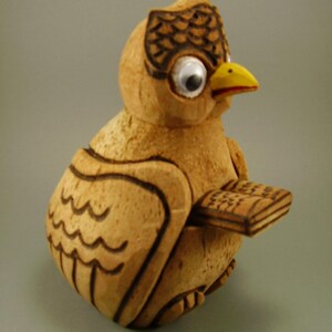 Könnte beinhalten: Eine geschnitzte Holzfigur eines Vogels, hellbraun mit dunkelbraunen geätzten Details, mit gelbem Schnabel und großen Wackelaugen. Der Vogel hält ein kleines, rechteckiges Objekt. Der Kopf hat ein dekoratives Muster.